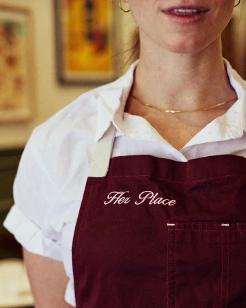 Close-up of a person wearing a white collared shirt and a burgundy apron with embroidered text, photographed by Alex Lau.
