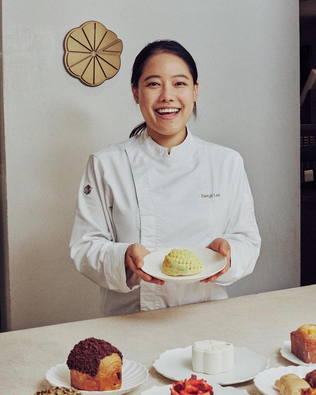 Smiling Asian chef in white coat presents pastry on plate in bright indoor scene with other desserts on table, neutral background and flower accent.