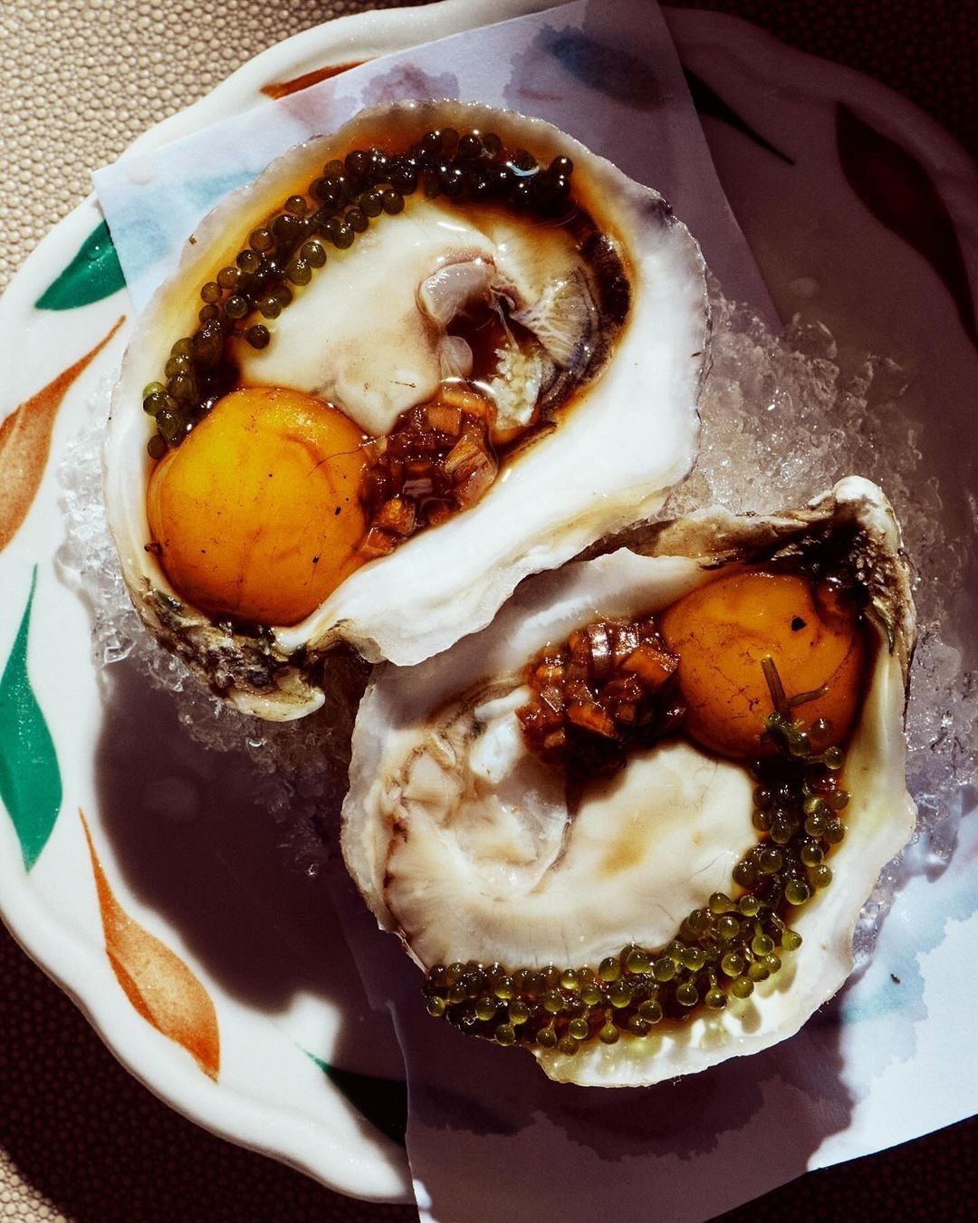 Overhead shot of two oysters on the half shell presented on ice with roe, served on a decorative plate with a white paper liner.