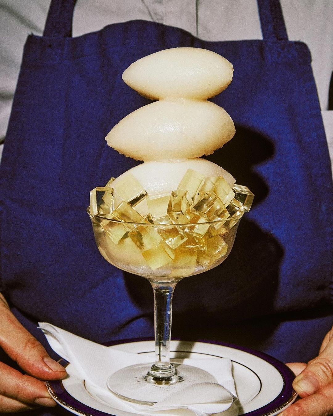 Elegant dessert presentation: Sorbet spheres atop jelly cubes in a stemmed glass, held on a plate with a white linen napkin, against a blue apron.