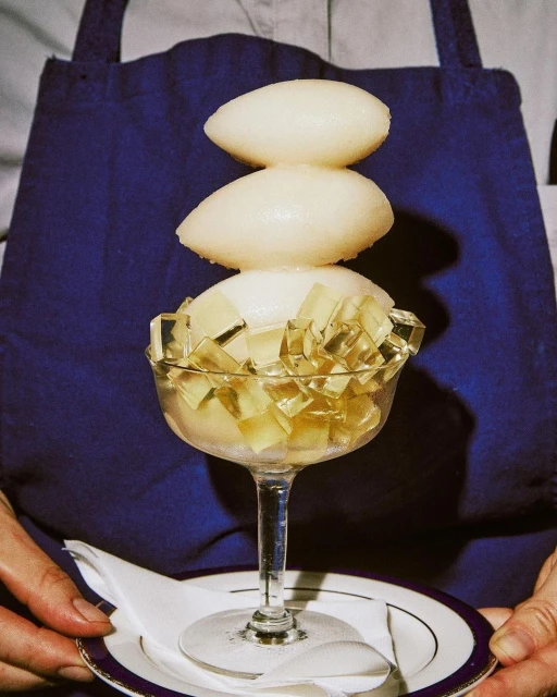 Elegant dessert presentation: Sorbet spheres atop jelly cubes in a stemmed glass, held on a plate with a white linen napkin, against a blue apron.