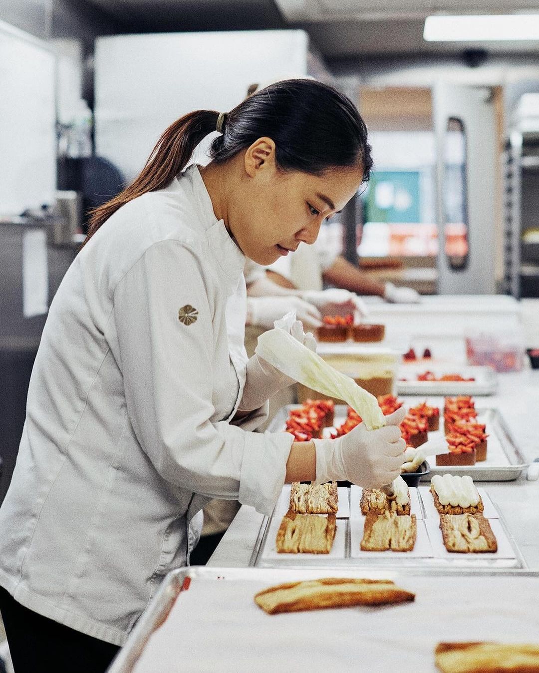 Pastry chef carefully decorates pastries with white icing in a bright commercial kitchen, showcasing baking process and culinary expertise.