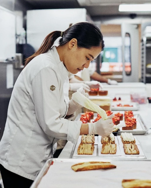Pastry chef carefully decorates pastries with white icing in a bright commercial kitchen, showcasing baking process and culinary expertise.