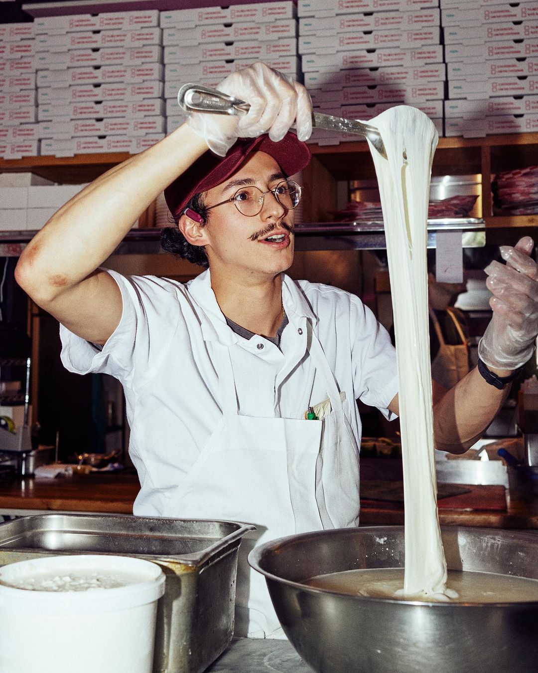 Person in kitchen pulling strands of dough, wearing white apron and cap, boxes in background, food preparation scene.