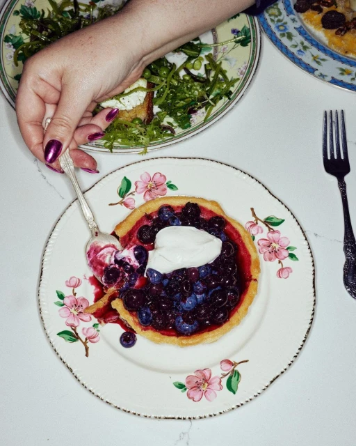 Overhead shot of a person using a spoon to serve a bite of blueberry tart topped with cream on a floral patterned plate.