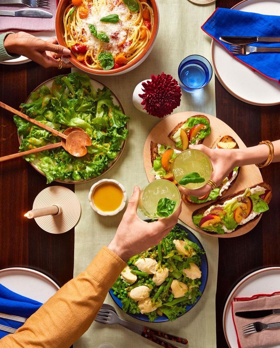 Overhead view of a dining table with plates of salad, pasta, bruschetta, and cocktails, featuring hands reaching for drinks and food.