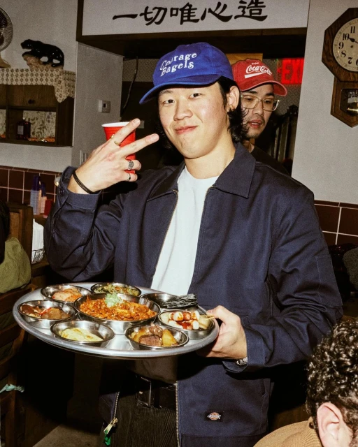 Man holding tray with Korean side dishes at indoor restaurant. He wears a blue cap, and another person is visible in the background.