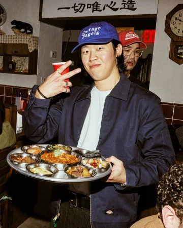 Man holding tray with Korean side dishes at indoor restaurant. He wears a blue cap, and another person is visible in the background.