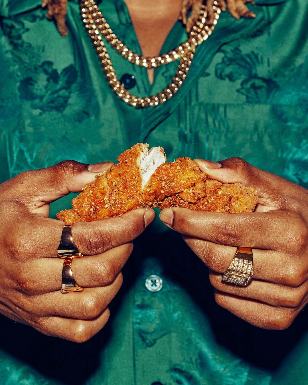 Close-up of a person holding a piece of fried chicken, wearing a green floral shirt and jewelry, showcasing a casual food presentation.
