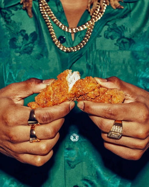 Close-up of a person holding a piece of fried chicken, wearing a green floral shirt and jewelry, showcasing a casual food presentation.