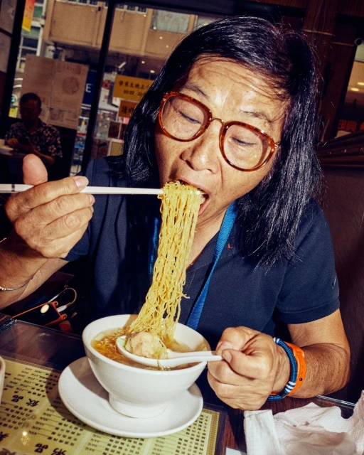 Woman with glasses enjoys a bowl of noodles with chopsticks at an indoor restaurant, with visible background activity.