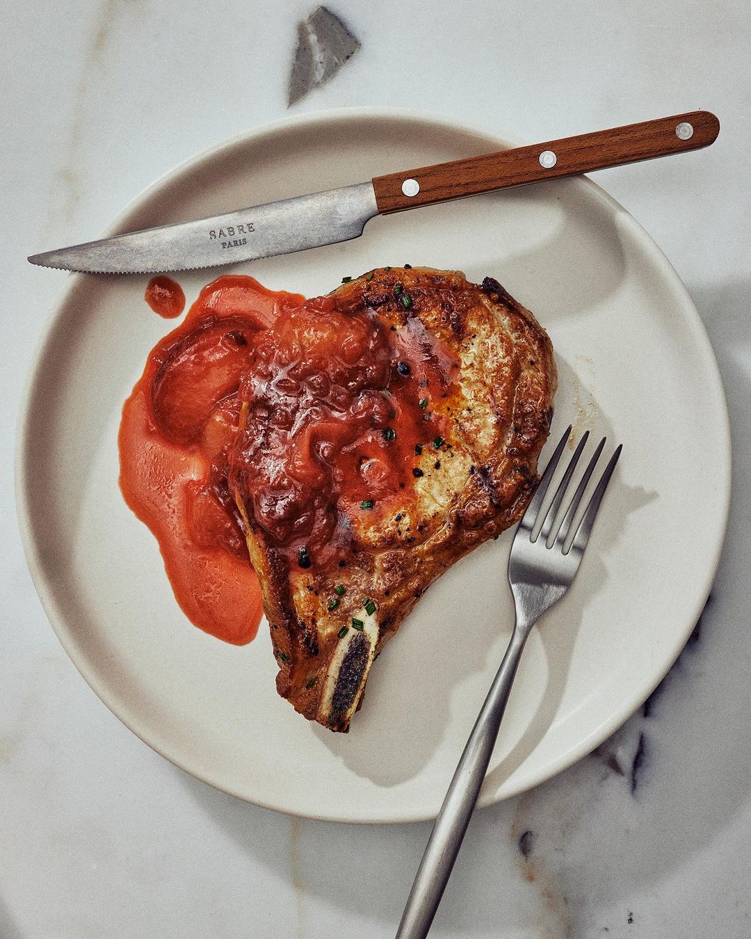 Overhead shot of a bone-in pork chop topped with red sauce on a white plate with a knife and fork on a marble surface.
