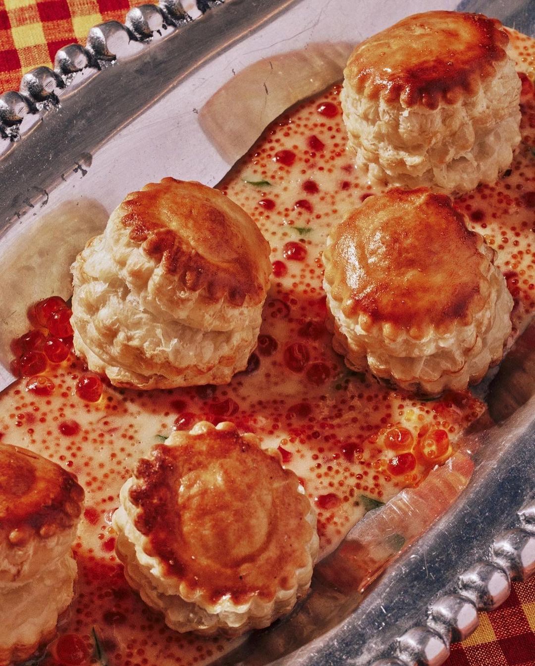 Elevated shot of golden vol-au-vents filled with creamy sauce and red caviar-like toppings on a silver platter, with a checkered cloth backdrop.