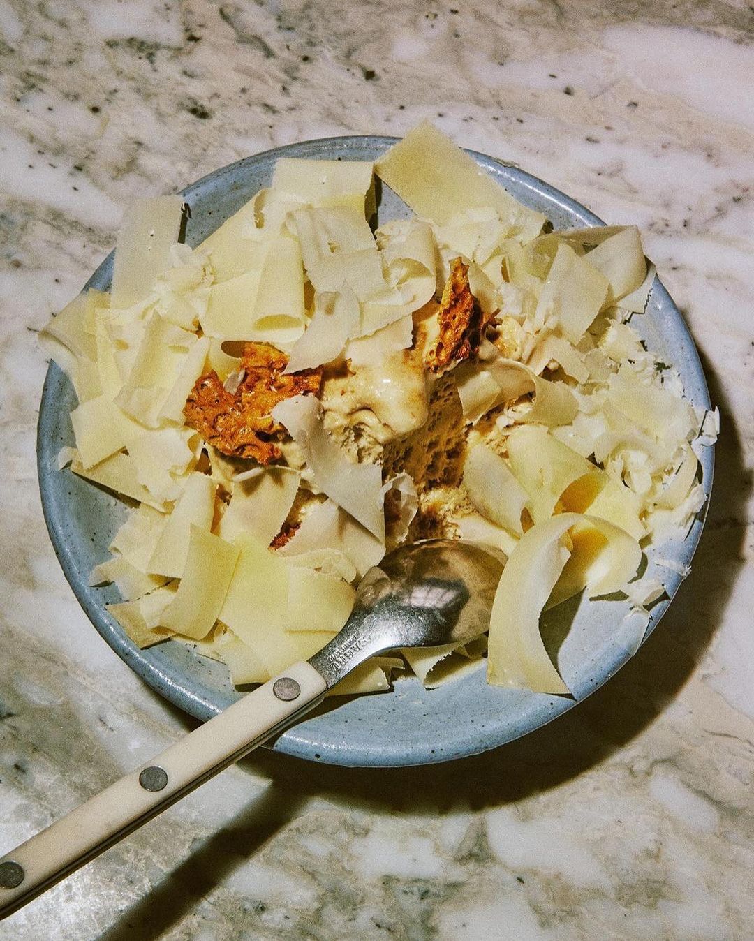 Overhead shot of a blue bowl filled with pasta topped with shavings of cheese and a spoonful of sauce, served with a silver spoon.
