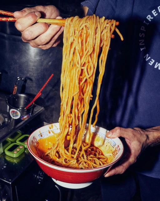 A person holding a bowl of ramen noodles high in the air with chopsticks, showcasing the noodles in a restaurant setting.
