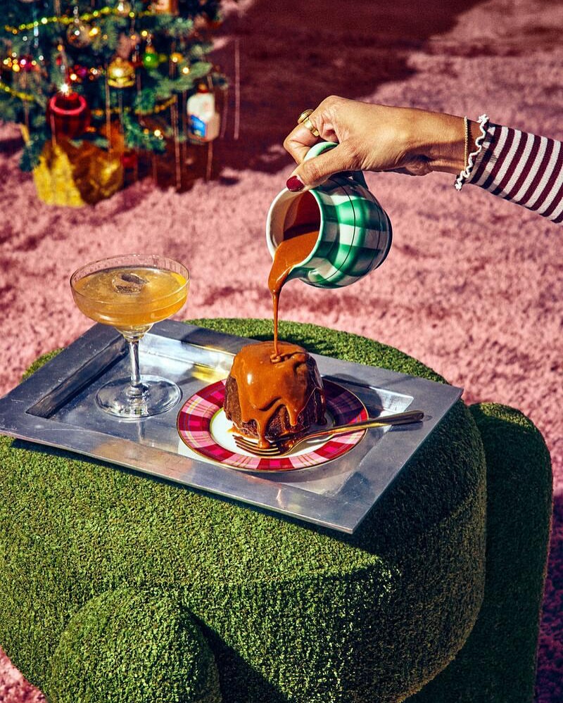Caramel sauce being poured onto a cake on a silver tray next to a cocktail, with a Christmas tree in the background.