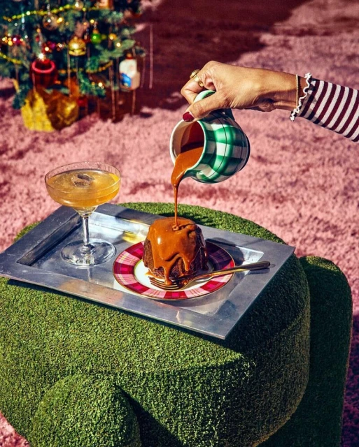 Caramel sauce being poured onto a cake on a silver tray next to a cocktail, with a Christmas tree in the background.