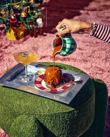 Caramel sauce being poured onto a cake on a silver tray next to a cocktail, with a Christmas tree in the background.
