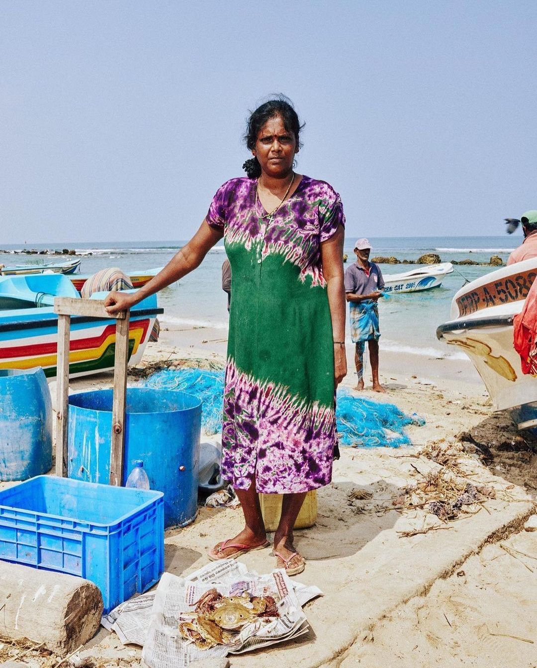 An Asian woman stands on a sandy beach next to fishing boats and gear. She wears a tie-dye dress and the sky is clear and sunny.