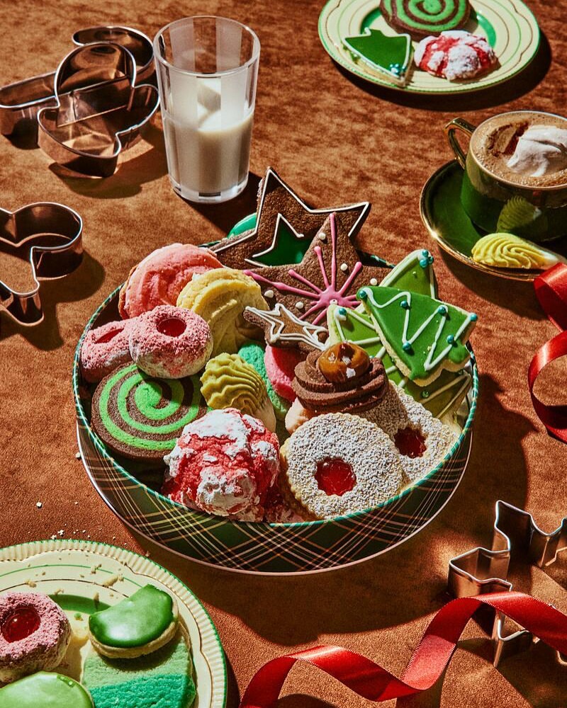 Festive holiday cookies arranged in a tin with a glass of milk, coffee cup, cookie cutters, and red ribbon on a brown surface.