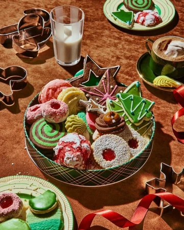 Festive holiday cookies arranged in a tin with a glass of milk, coffee cup, cookie cutters, and red ribbon on a brown surface.