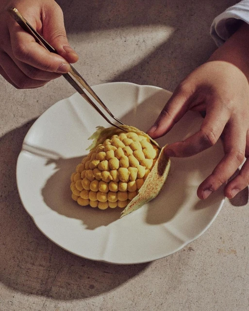 Overhead shot of a person using tweezers to peel back the husk on a corn-shaped dessert on a white plate, casting shadows.