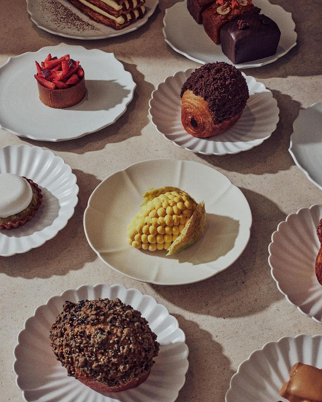 Overhead shot of an assortment of miniature cakes and pastries on decorative white plates, arranged on a neutral-toned surface with soft lighting.