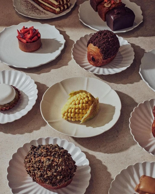 Overhead shot of an assortment of miniature cakes and pastries on decorative white plates, arranged on a neutral-toned surface with soft lighting.