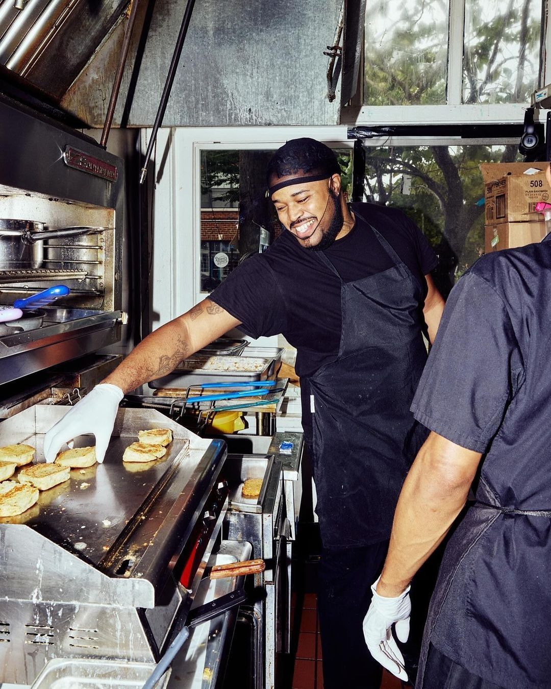 A smiling man in a black apron cooking biscuits on a flat top grill in a commercial kitchen with stainless steel equipment and natural light.
