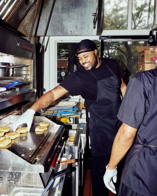 A smiling man in a black apron cooking biscuits on a flat top grill in a commercial kitchen with stainless steel equipment and natural light.