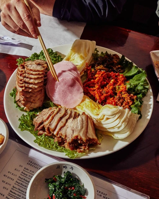 Overhead shot of white plate piled high with Korean braised pork, kimchi, lettuce, cabbage and ham, with chopsticks picking at food.