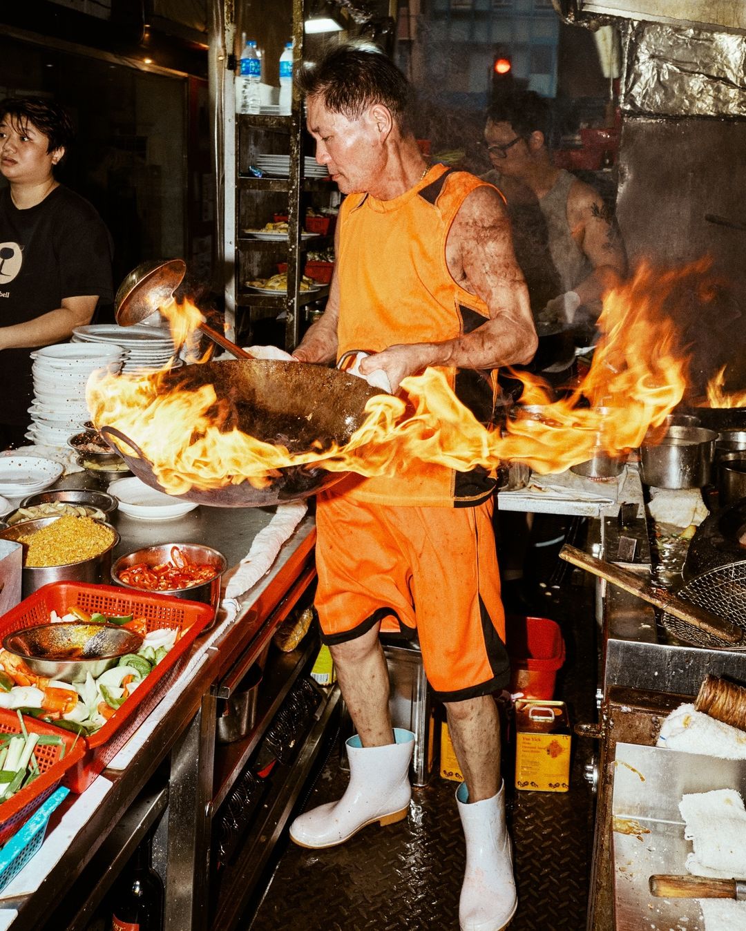An adult male chef in an orange uniform expertly cooks with a wok engulfed in flames in a professional restaurant kitchen setting.