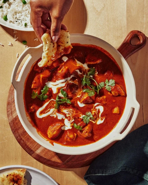 Overhead shot of a bowl of butter chicken topped with cream and cilantro, served with rice and bread on a wooden surface.