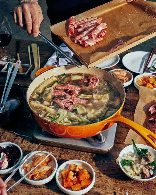 Top-down view of a person adding raw meat to a Korean hot pot on a wooden table with various Korean side dishes around it.