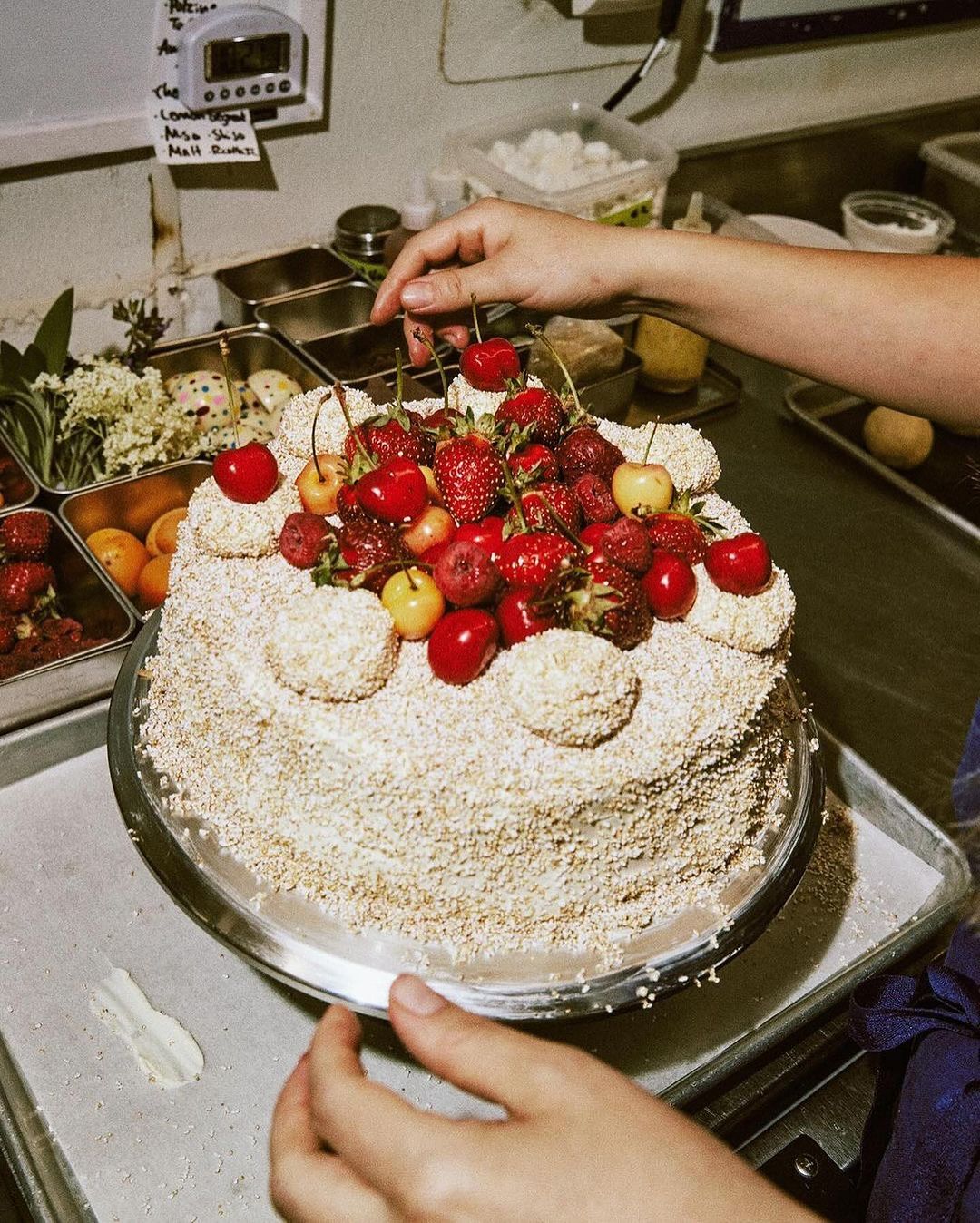 Chef decorating a frosted cake with fresh strawberries and cherries in a commercial kitchen, showcasing culinary artistry and dessert preparation.