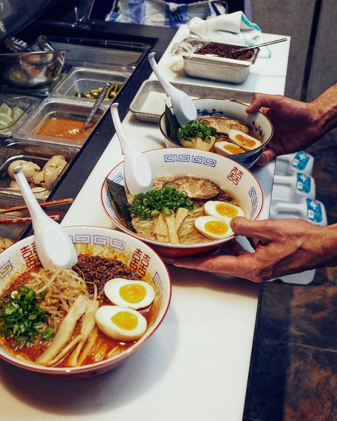 Three bowls of ramen with eggs and green onions are held by a person, with ingredient containers visible in the food service area.