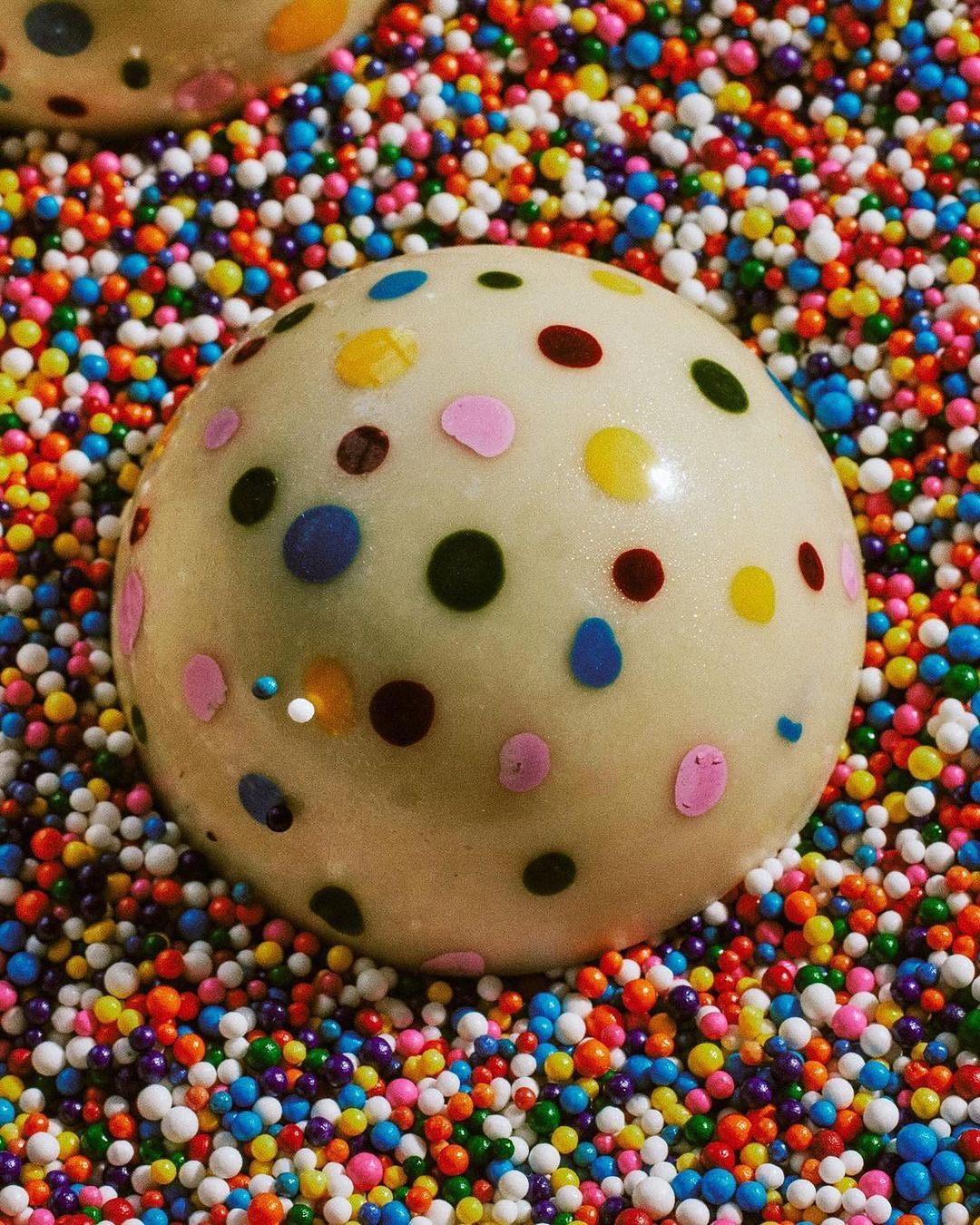 Close-up of a candy ball covered in colorful dots, sitting on a bed of multi-colored sprinkles. Food photography showcases a sugary treat.