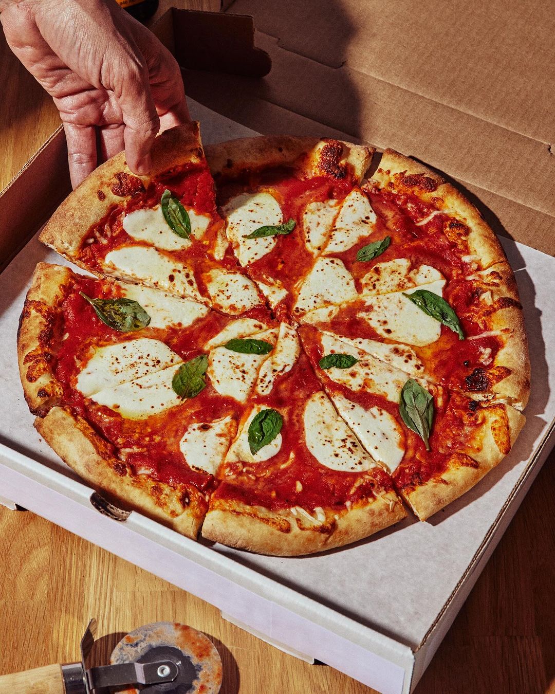 Overhead shot of a fresh Margherita pizza in a white box, ready to eat, with a pizza cutter and a hand reaching for a slice.