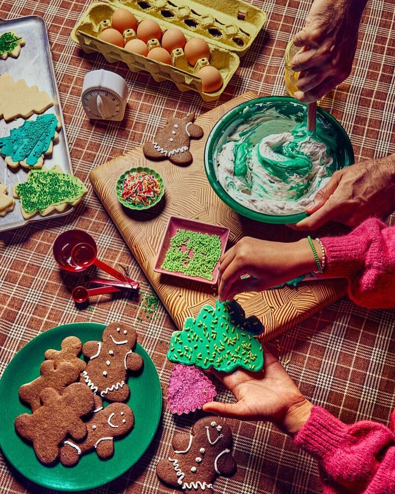 Overhead shot of people decorating Christmas cookies on a checkered tablecloth, featuring cookies, eggs, frosting, and baking tools.