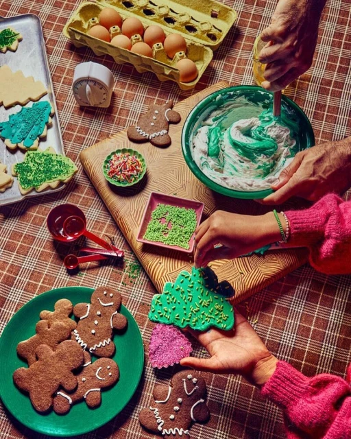 Overhead shot of people decorating Christmas cookies on a checkered tablecloth, featuring cookies, eggs, frosting, and baking tools.