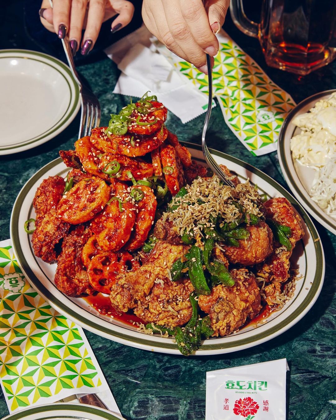 Overhead shot of an oval plate piled high with Korean fried chicken and lotus root, set on a green marble table with patterned napkins.