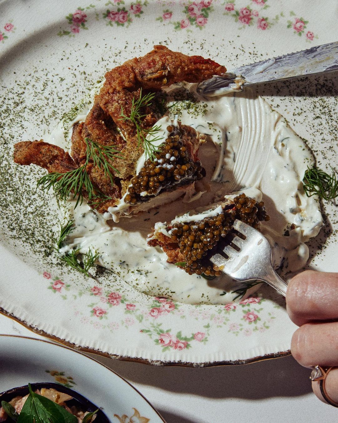 Overhead shot of fried food topped with caviar and creamy sauce on a floral patterned plate with a fork and hand holding it.