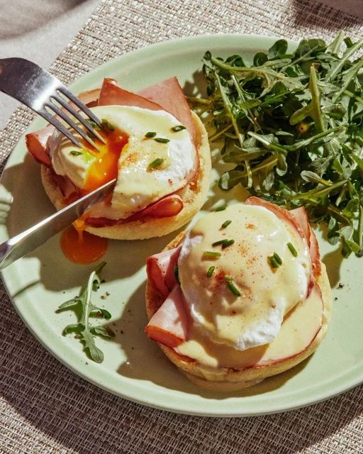Eggs Benedict with ham and hollandaise sauce on an English muffin, served with a side of arugula salad, being cut by a fork and knife.