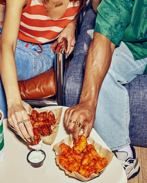 Two people sharing platters of chicken wings, seated on a couch, enjoying a casual meal with dipping sauce and a beverage on a table.