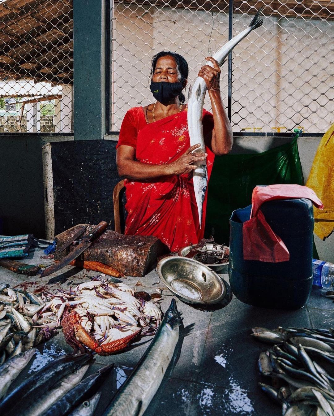 A woman in a red dress and face mask stands in an indoor market holding a large fish with various smaller fish on the counter.