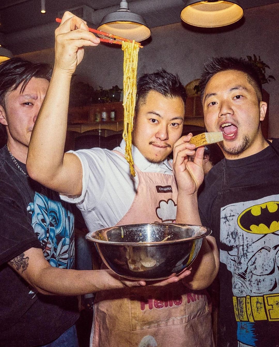 Three Asian men pose indoors with food. One holds noodles with chopsticks, another a metal bowl, and another holds a utensil to mouth.