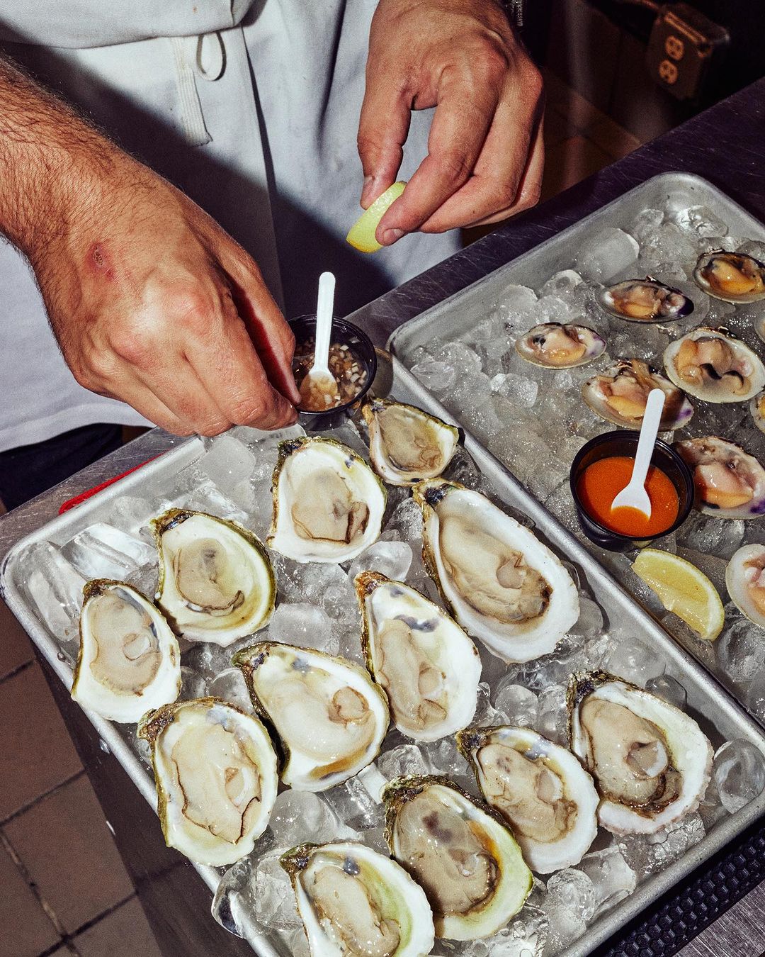 Overhead shot showcases a tray of fresh oysters on ice alongside clams, with an adult hand squeezing lemon, highlighting food photography.