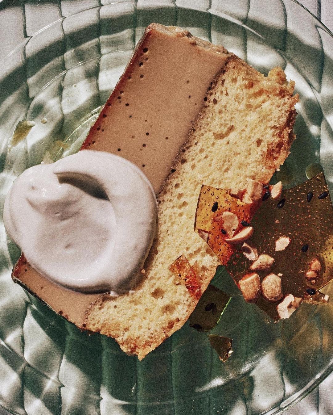 Overhead shot of a slice of flan cake on a glass plate, topped with a dollop of cream and garnished with nuts and brittle.