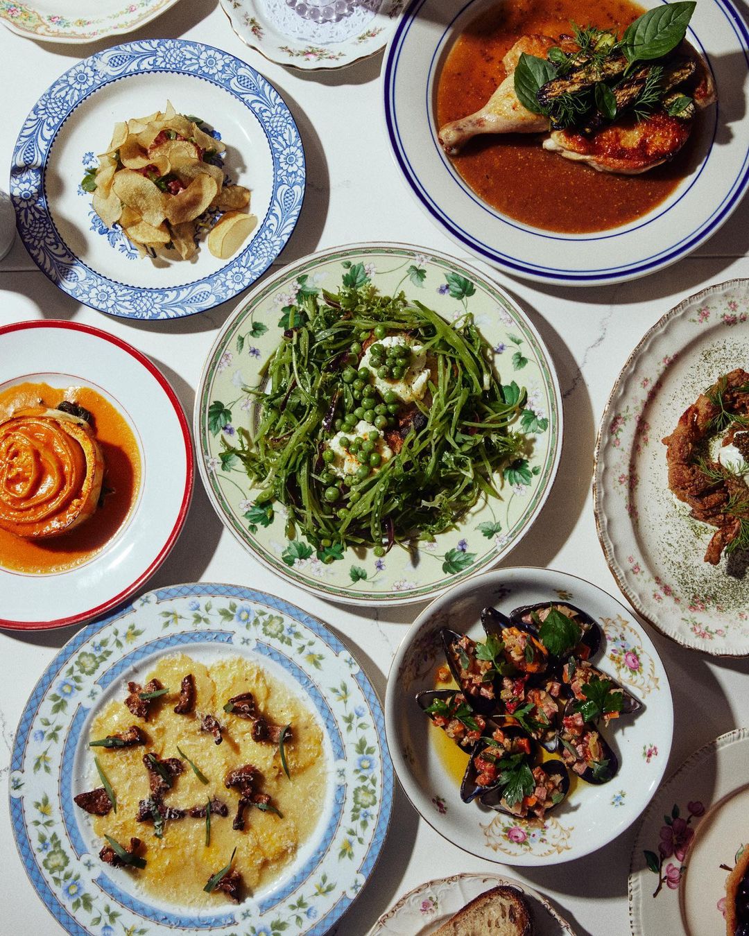 Overhead shot of beautifully arranged dishes on patterned plates featuring chicken, greens, mussels, and pastries on a white table.