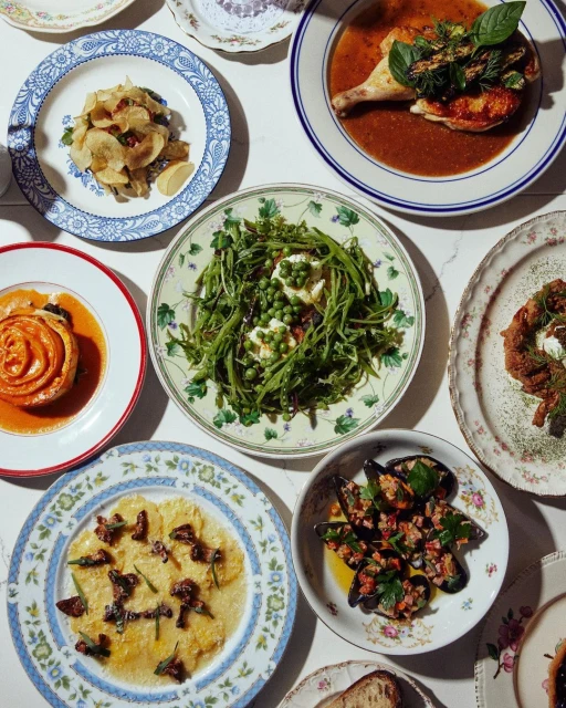 Overhead shot of beautifully arranged dishes on patterned plates featuring chicken, greens, mussels, and pastries on a white table.